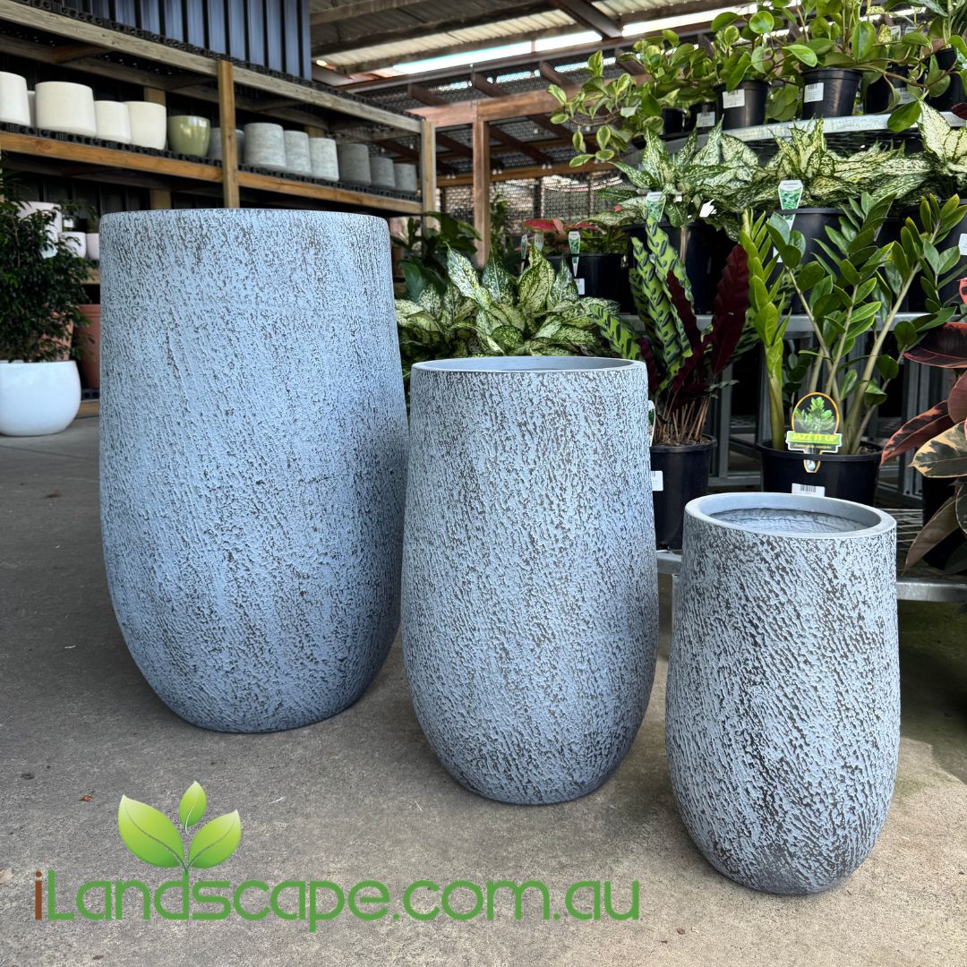 Three gray stone-textured planters in a garden center setting with plants and shelves in the background.