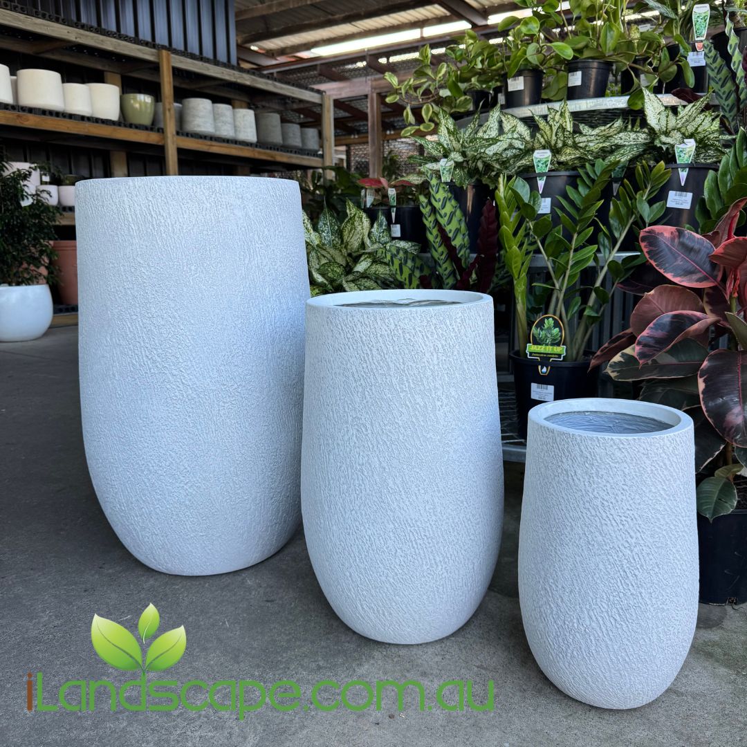 Three textured white planters in a garden center setting with plants and shelves in the background.